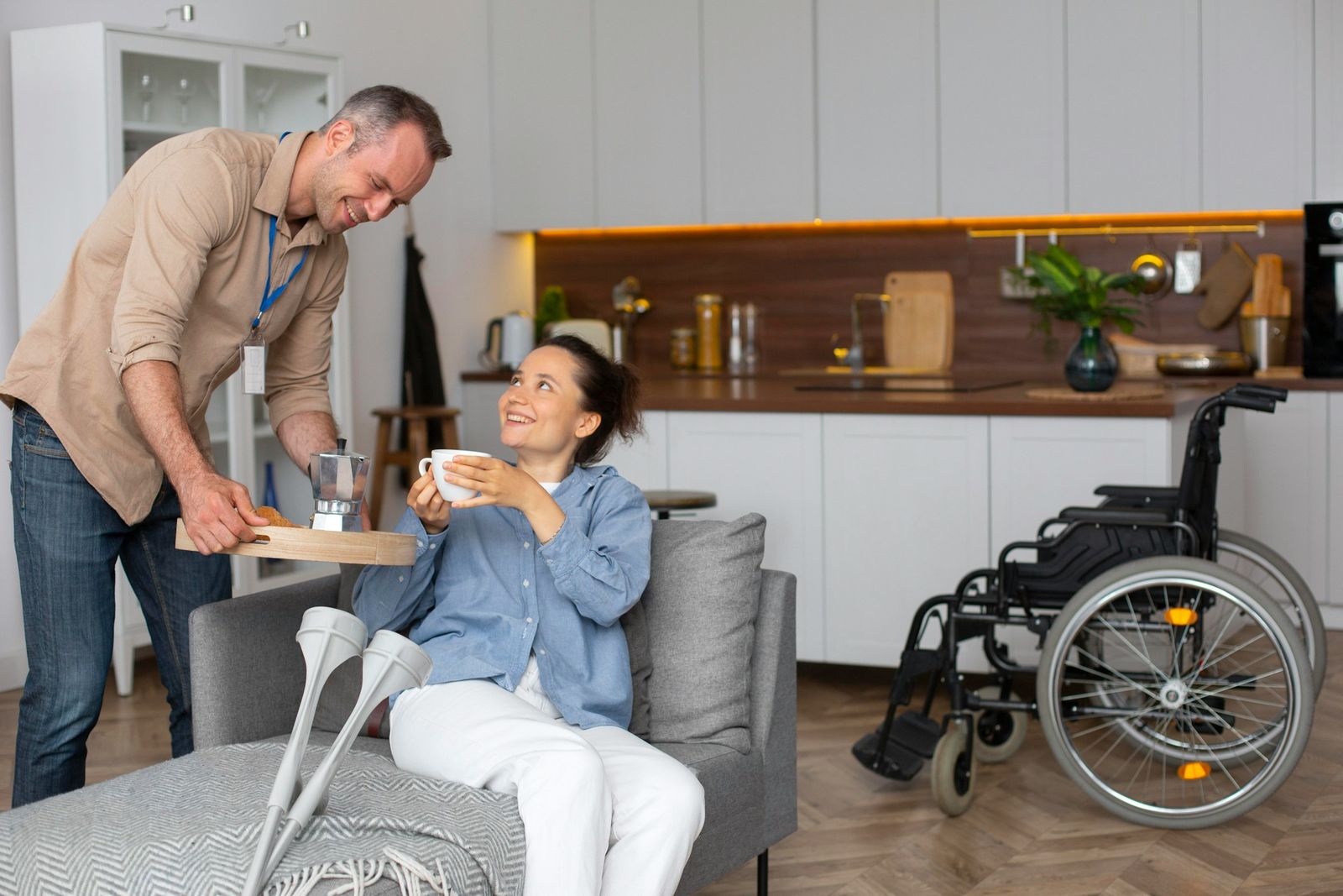 Dans un salon, un intervenant apporte une tasse à une jeune femme assise sur le canapé, ses béquilles à côté d'elle et un fauteuil roulant en arrière-plan ; ils échangent un sourire.