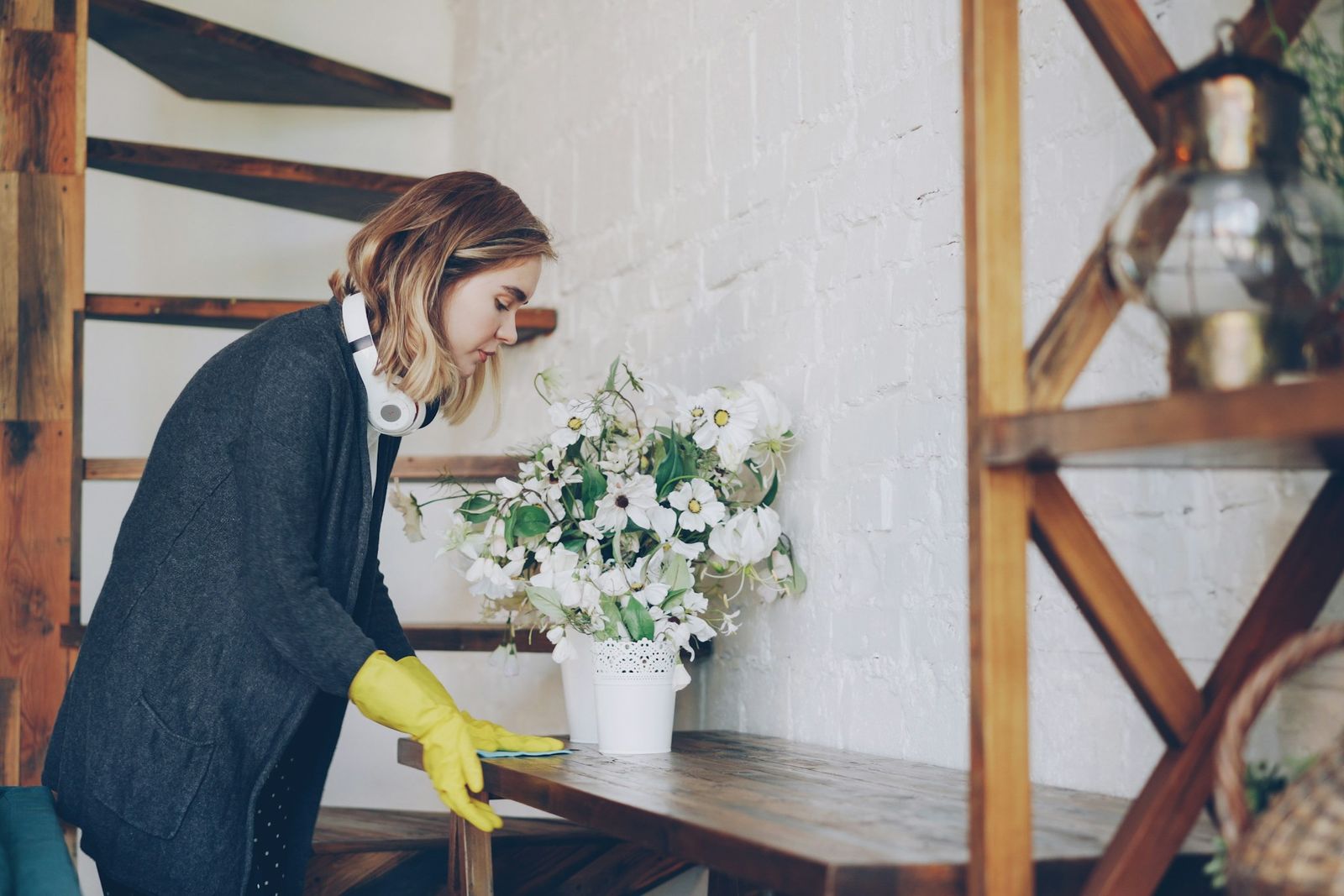 Une femme en gants de ménage nettoie une console près d'un bouquet de fleurs, dans un intérieur chaleureux.